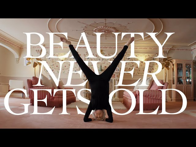 A senior woman does a handstand in a traditionally-decorated living room. Text behind her reads: "Beauty Never Gets Old."