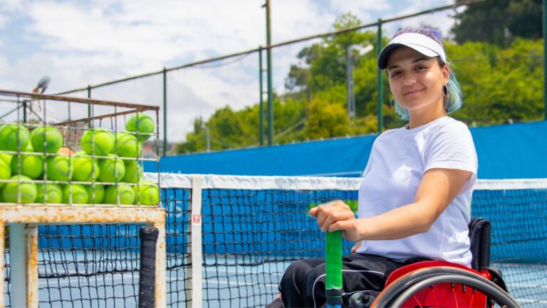 A female wheelchair user on the tennis court, wearing a sun hat and smiling.