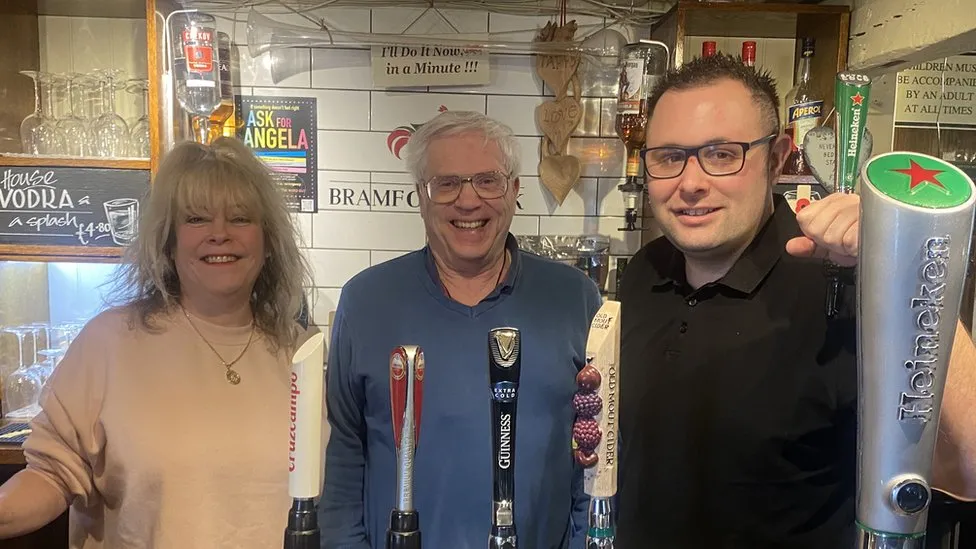 The owners of The Bramford Cock smiling next to David, a white man with short white hair and glasses, their hearing impaired customer providing BSL classes at the pub.