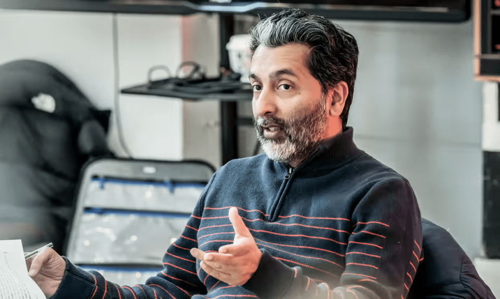 A picture of Amit Sharma, an Indian man with short black and grey hair and beard, wearing a navy blue jumper with red stripes.