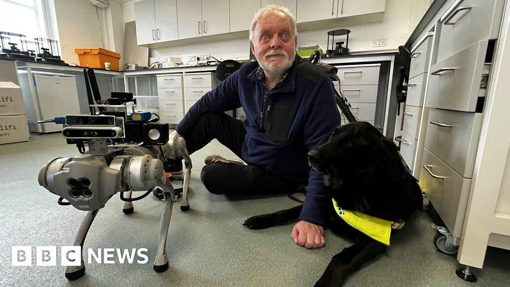 A blind man is sitting on the floor next to his guided dog and the robot dog prototype.