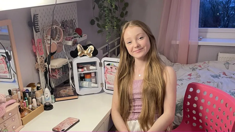 A teenager is smiling in their room, next to their desk full of skincare products.