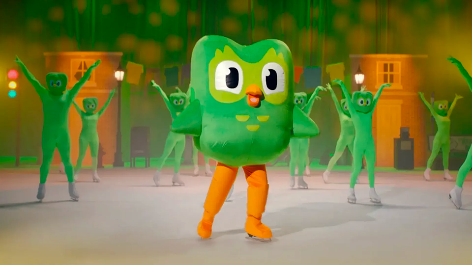 The Duolingo bird is ice skating with a group of backup dancers in a fake performance.
