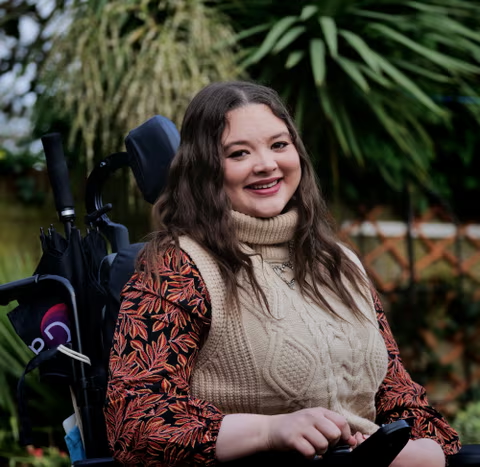 A picture of Amy, a white woman with long brown hair, wearing an autumnal outfit. She's smiling at the camera while sitting on her powered wheelchair.