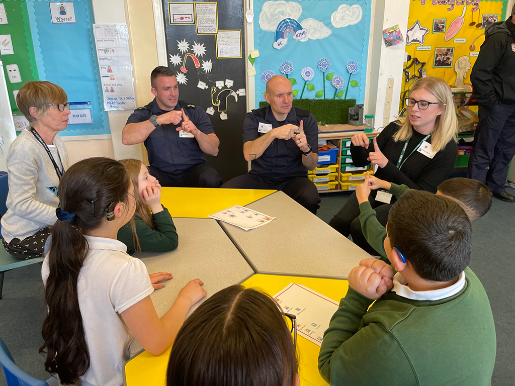 A group of deaf students sit around a school table teaching some firefighters some sign language.