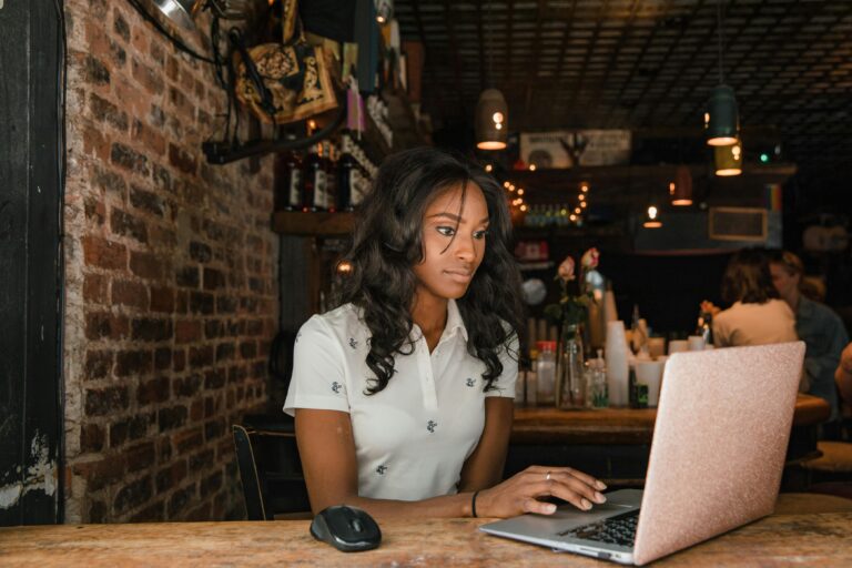 A Black woman with an upper limb difference is in a coffee shop looking at her laptop.