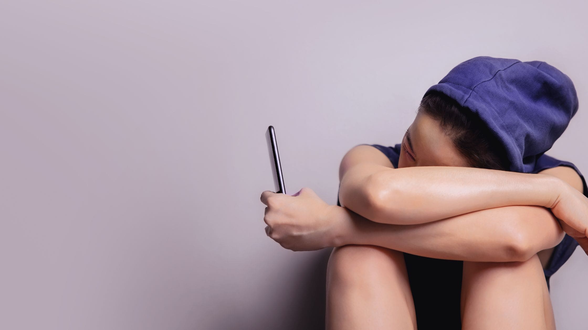 Woman in a hoodie sat on the floor with her head in her arms scrolling social media on her phone - sugesting she needs a more healthy relationship with social media.