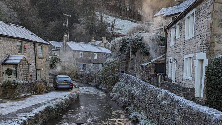 An image from Castleton, a village in the Peak District, showing some rural houses and a river in the middle, everything covered in snow. 