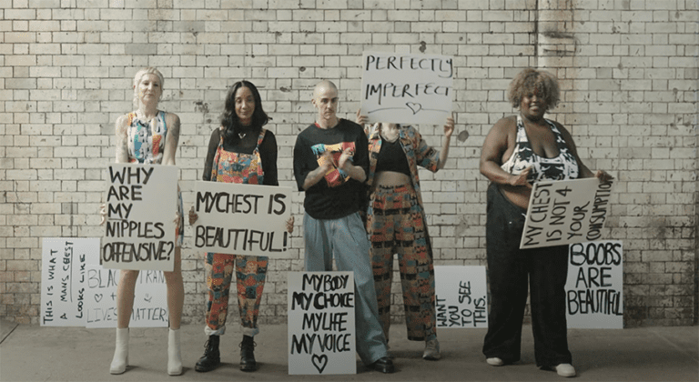 A group of five people, including women, a non-binary person and a trans man stand against an indoor brick wall. They are holding signs, as well as there being some more against the wall in the background. These signs have phrases on them such as "My chest is beautiful," and "Why are my nipples offensive?"