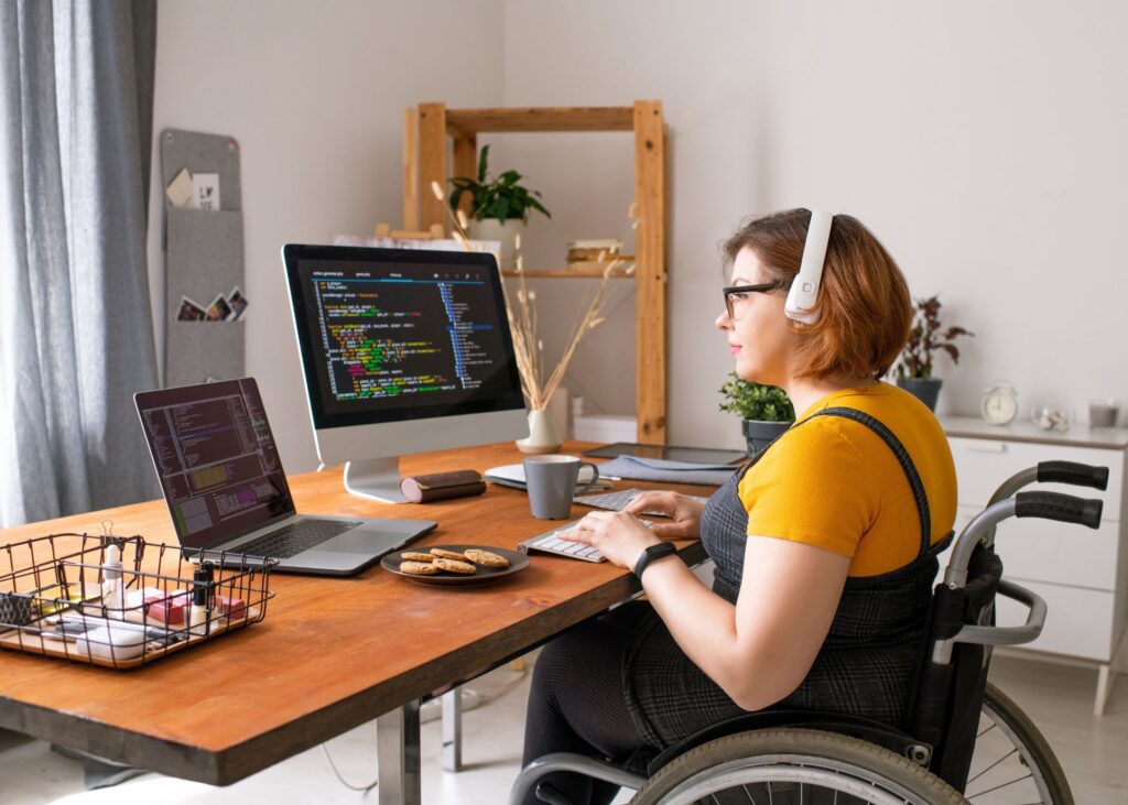 A female wheelchair user is wearing headphones while coding on her home computer setup.